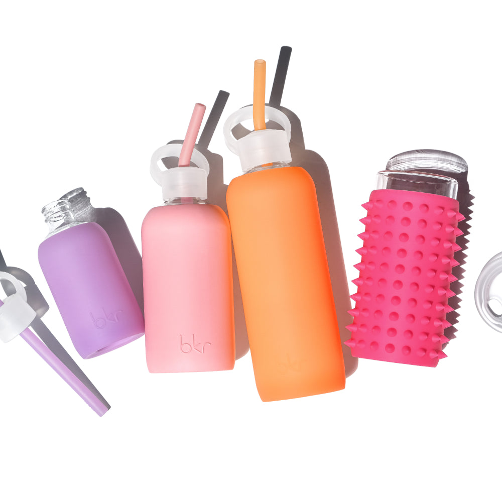 Reusable glass and silicone water bottles in sheer orange cream soda, opaque pastel fluorescent coral, and sheer orchid violet.